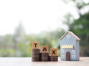 A model home sits next to stacks of coins with wooden blocks spelling out the word “Tax” on top.