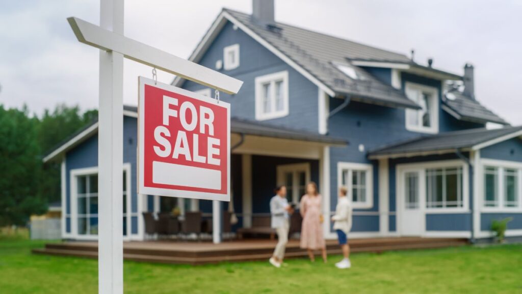 A blue-grey two story home on a beautiful green lawn, out-of-focus, with focus and emphasis on the For Sale sign