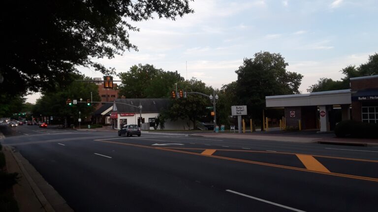 Wide multi-lane Broad Street (Route 7) in Falls Church VA with a turn lane, traffic signals, low commercial buildings and mature shade trees under a partly cloudy sky