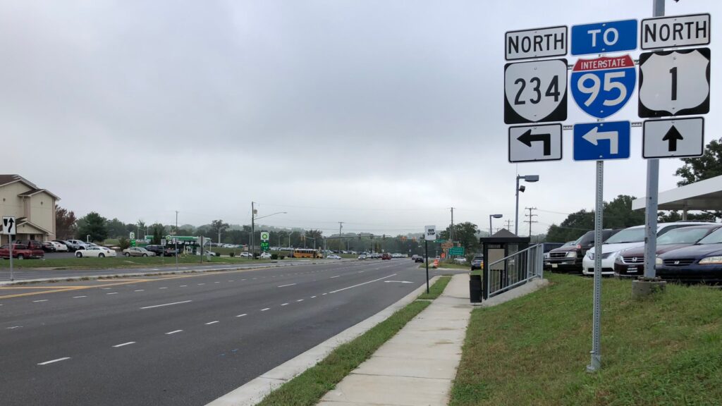 Multi-lane U.S. Route 1 heading north through Dumfries VA, with green signs for Route 234 and Interstate 95, a sidewalk and grass strip, and businesses along the road