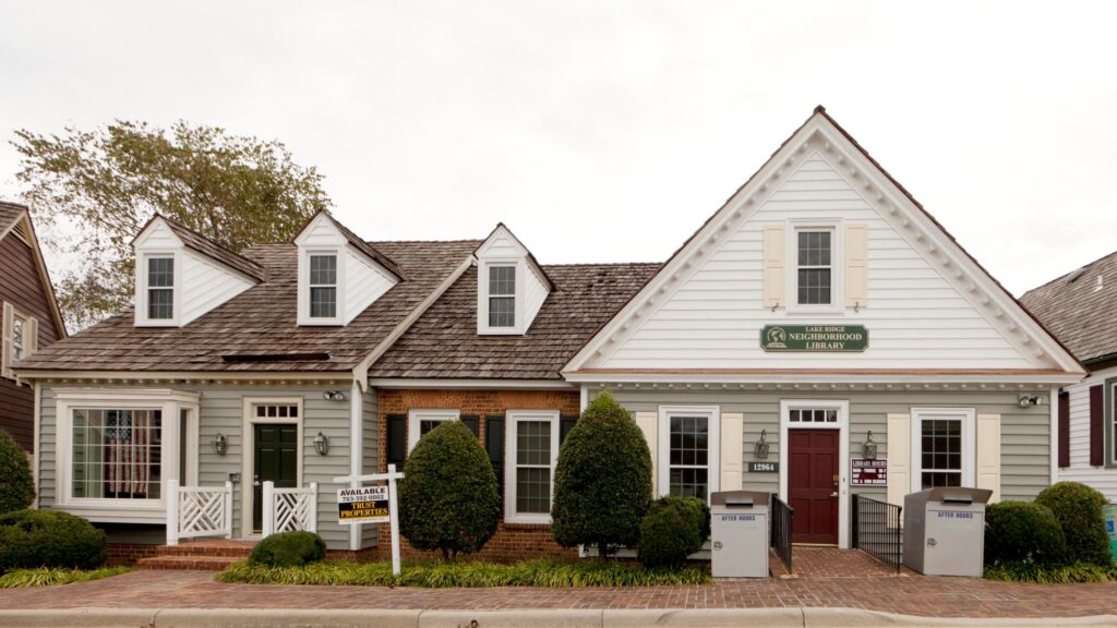 Two-story gray and white building with dormer windows, a red door and landscaped shrubs, home to the Lake Ridge Library next to Tackett’s Mill in Lake Ridge VA