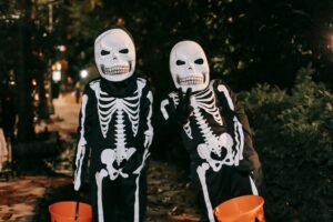 Two children in skeleton costumes holding orange pumpkin buckets while trick-or-treating on a neighborhood sidewalk at dusk