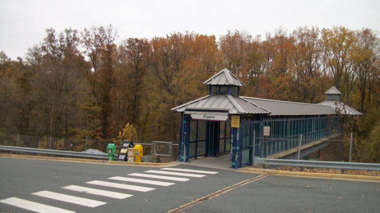 Covered pedestrian footbridge with a Rippon sign and peaked roof at the Rippon VRE station in Woodbridge VA, with a crosswalk in front and autumn trees behind