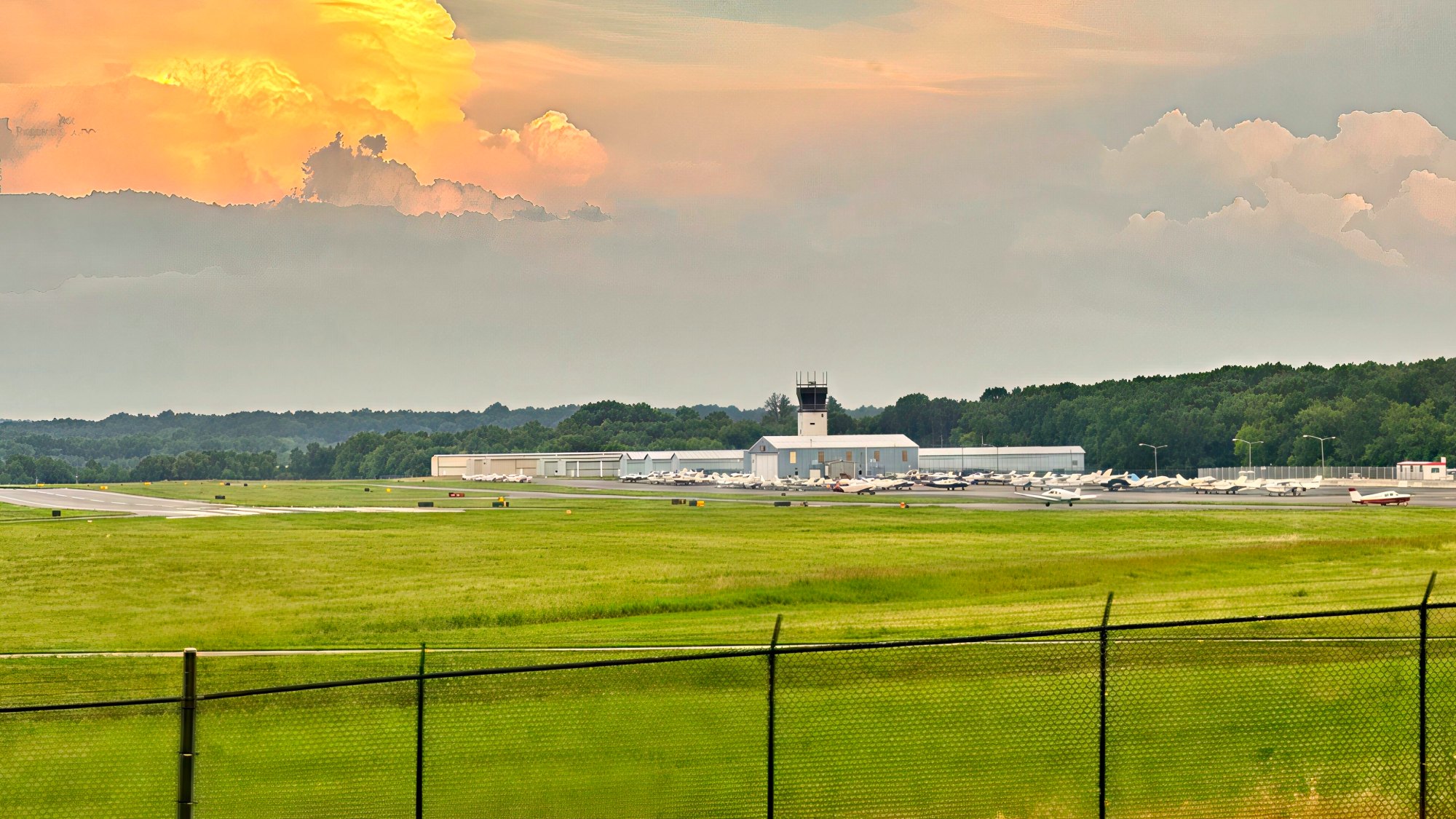 Wide grass airfield behind a chain-link fence with the control tower and low terminal buildings at Washington Manassas Airport in Manassas VA under an orange evening sky